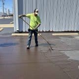 Two workers are resurfacing a parking lot, undertaking crucial asphalt maintenance. One worker, in a bright yellow shirt and cap, is spreading a brown sealant with a long-handled tool, while the other stands nearby. They work beside a large metal building.