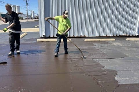 Two workers are resurfacing a parking lot, undertaking crucial asphalt maintenance. One worker, in a bright yellow shirt and cap, is spreading a brown sealant with a long-handled tool, while the other stands nearby. They work beside a large metal building.