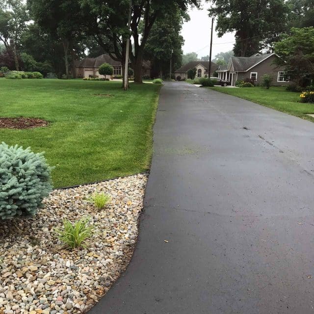 A freshly paved driveway, showcasing expert asphalt paving, curves through a suburban neighborhood, bordered by neatly trimmed grass and small landscaped areas with rocks and plants. Trees and houses line the background under an overcast sky.