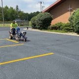 A person operates a line striping machine to paint yellow parking lines on a newly paved lot, ensuring precise asphalt maintenance. In the background, a brick building with a cross and green bushes stands under a partly cloudy sky.