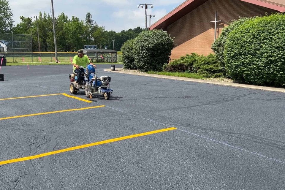 A person operates a line striping machine to paint yellow parking lines on a newly paved lot, ensuring precise asphalt maintenance. In the background, a brick building with a cross and green bushes stands under a partly cloudy sky.