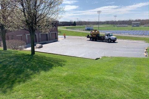 A truck is parked on a paved area near a grassy field and a sports track, possibly for some asphalt maintenance. A person in orange stands beside the truck. The sky is partly cloudy, and a tree with budding flowers graces the foreground.