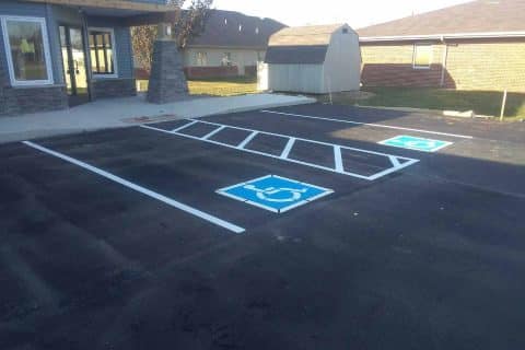 A parking lot features two marked handicap spaces in front of a building, outlined in blue with wheelchair symbols painted on the freshly maintained asphalt. Nearby, a small shed and two brick buildings are visible, demonstrating excellent asphalt maintenance practices.