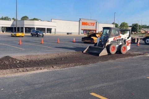 A construction vehicle tackles asphalt maintenance on a parking lot near an empty Big Lots store. Traffic cones line the area under repair, and two cars are parked in the background. The weather is clear and sunny, perfect for paving work.