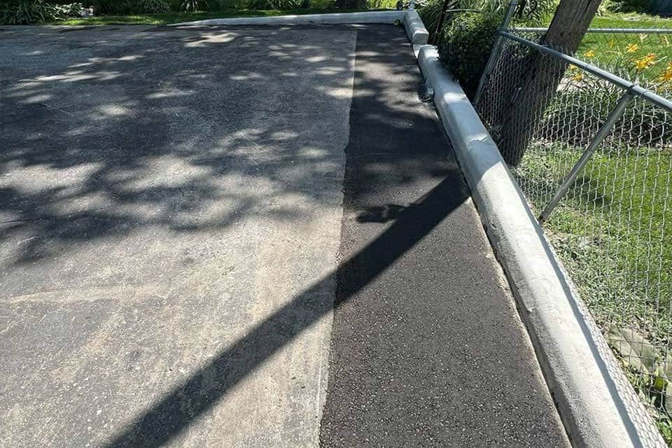 Paved road with asphalt repair beside a curb and chain-link fence.