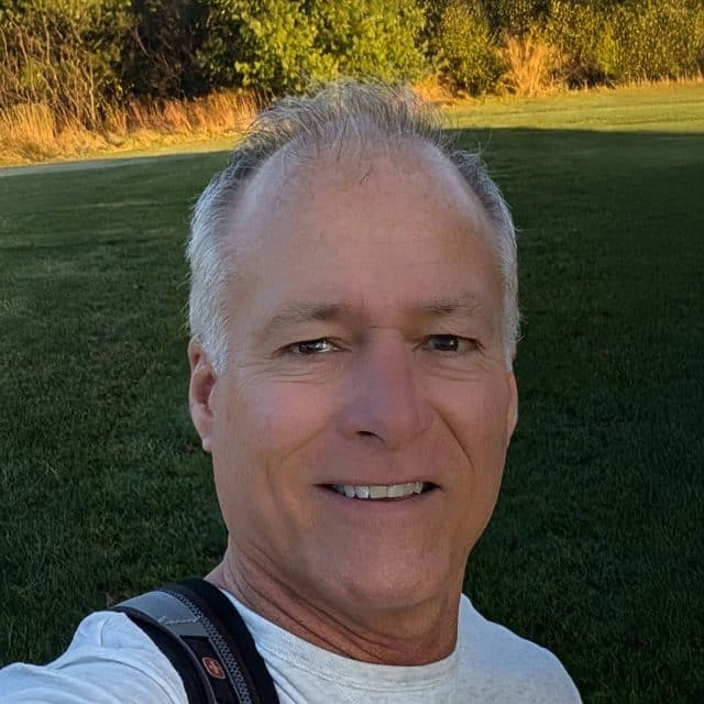 Man smiling outdoors in a grassy area with trees in the background.