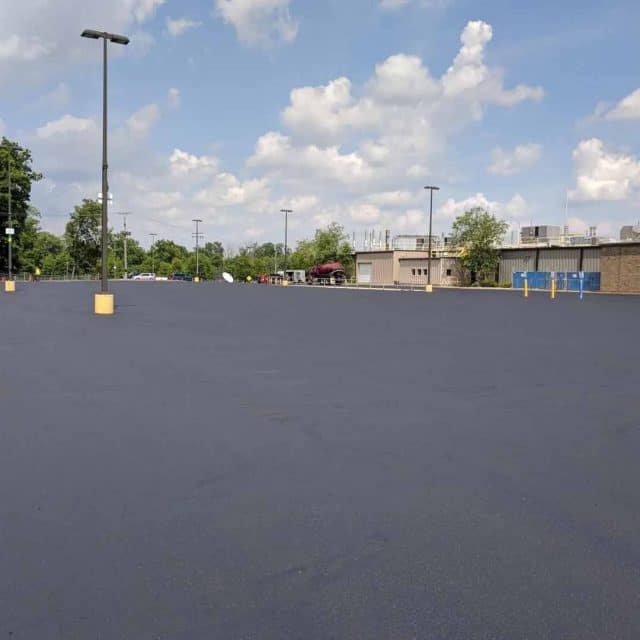 Empty parking lot with light poles and a distant building under a cloudy sky.