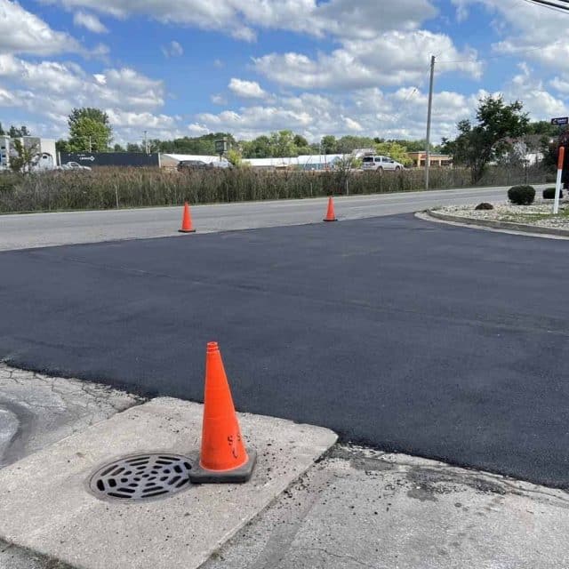 Freshly paved asphalt section with orange cones around it near a roadway.