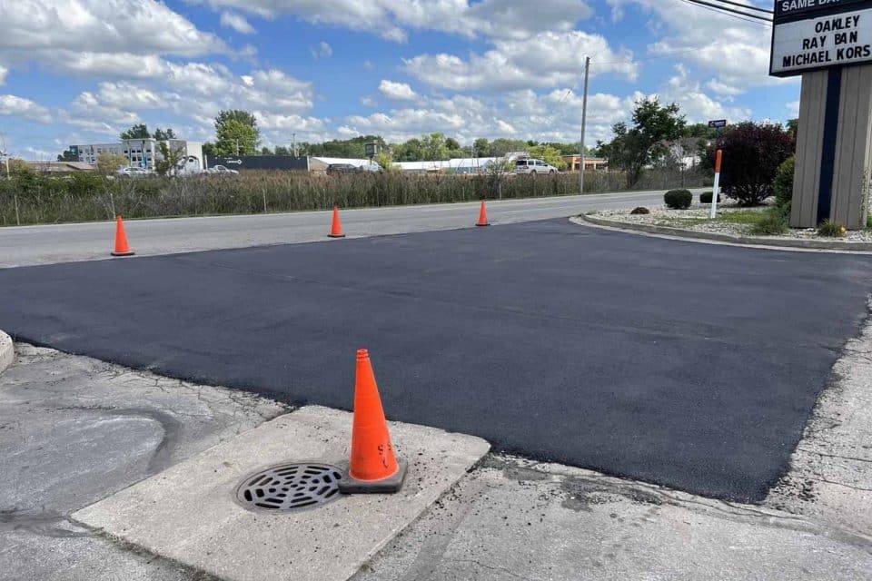 Freshly paved asphalt section with orange cones around it near a roadway.