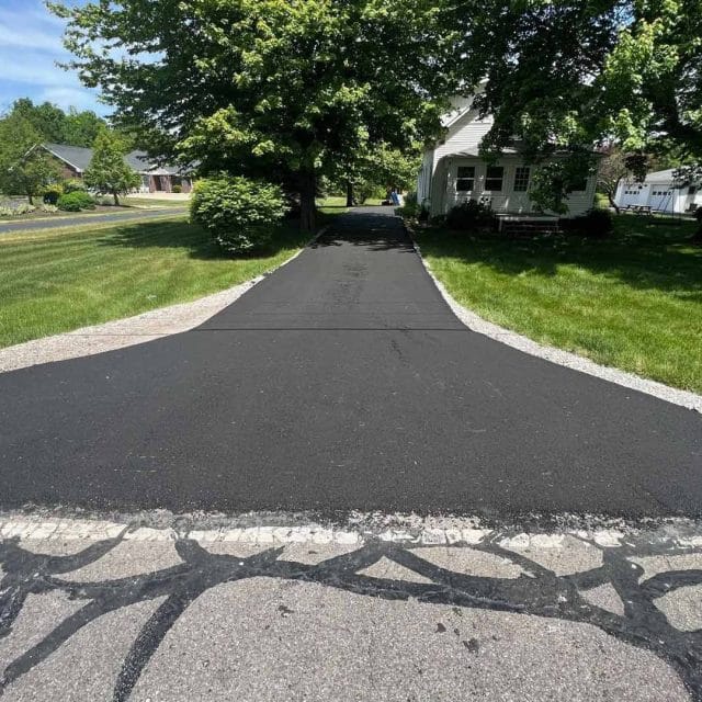 Freshly paved black driveway leading to a house surrounded by trees.