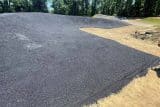 Freshly paved asphalt surface with surrounding dirt and trees in the background.