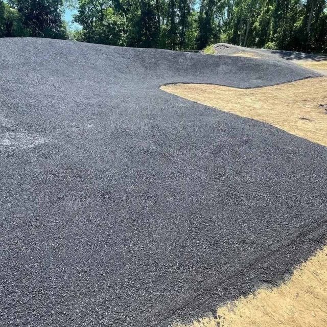 Freshly paved asphalt surface with surrounding dirt and trees in the background.