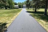 A newly paved asphalt driveway leads to a house with a garage, surrounded by trees.