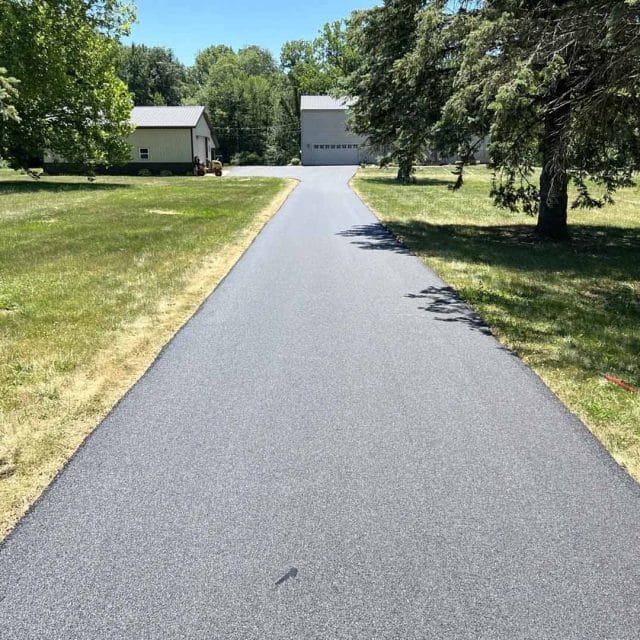 A newly paved asphalt driveway leads to a house with a garage, surrounded by trees.