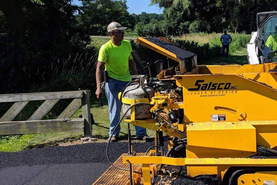 A worker in a neon yellow shirt stands beside a Salsco paving machine, expertly performing asphalt maintenance on a path in a wooded area. Another person is visible in the background near the wooden fence.