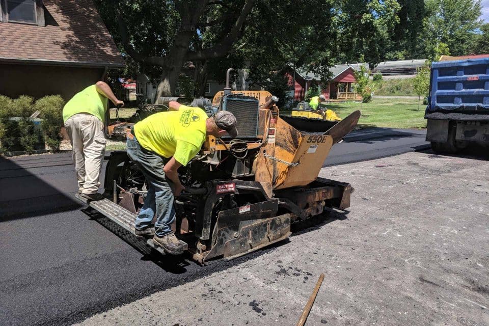 Workers in bright yellow shirts expertly manage an asphalt paving machine, laying fresh asphalt on a driveway. Nearby, a blue truck rests as trees and a red building enhance the sunny backdrop.