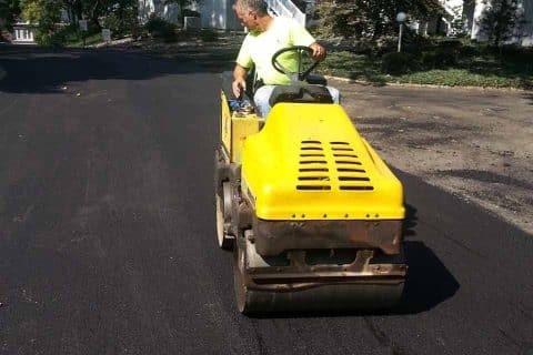 Person operating a small yellow road roller on freshly paved asphalt.