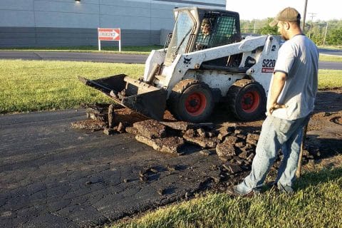 A skid steer loader breaks up asphalt while a worker observes.