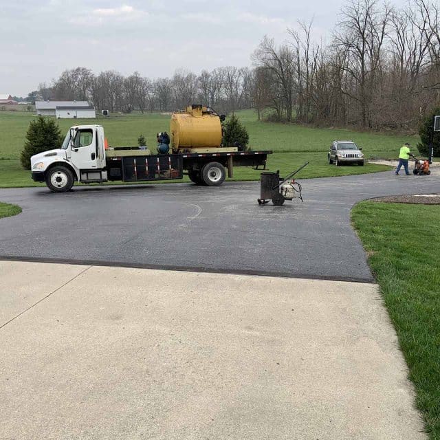 Truck and workers on a driveway, with a field and trees in the background.