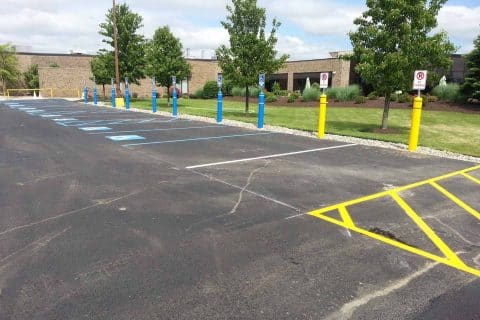 A newly paved parking lot, highlighted by recent asphalt paving, features designated accessible parking spaces marked in blue. Yellow stripes mark a no-parking zone. Surrounding landscaping includes green grass, trees, and several shrubs. A brick building is visible in the background.