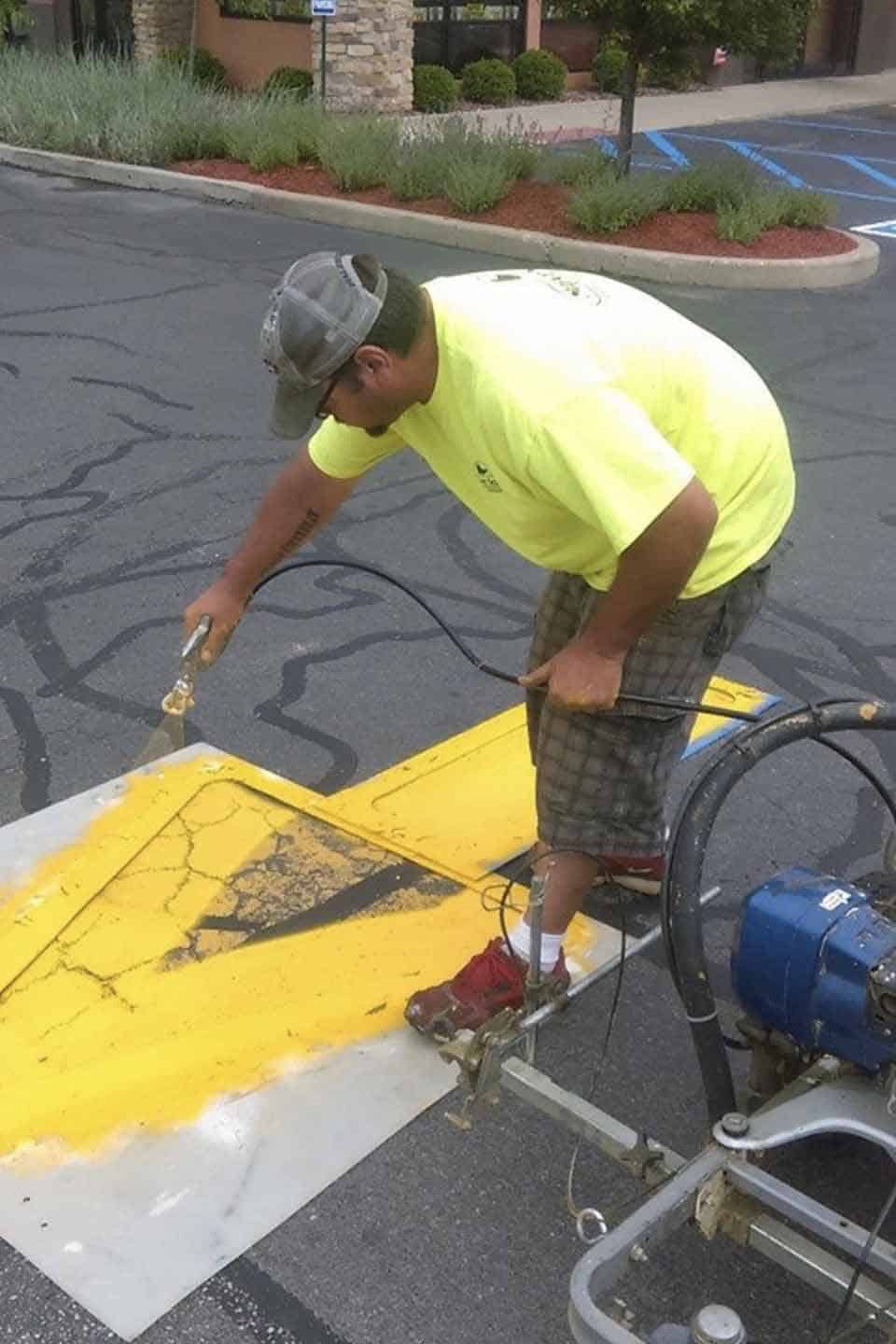 Worker spray-painting a yellow arrow on parking lot pavement with a stencil.